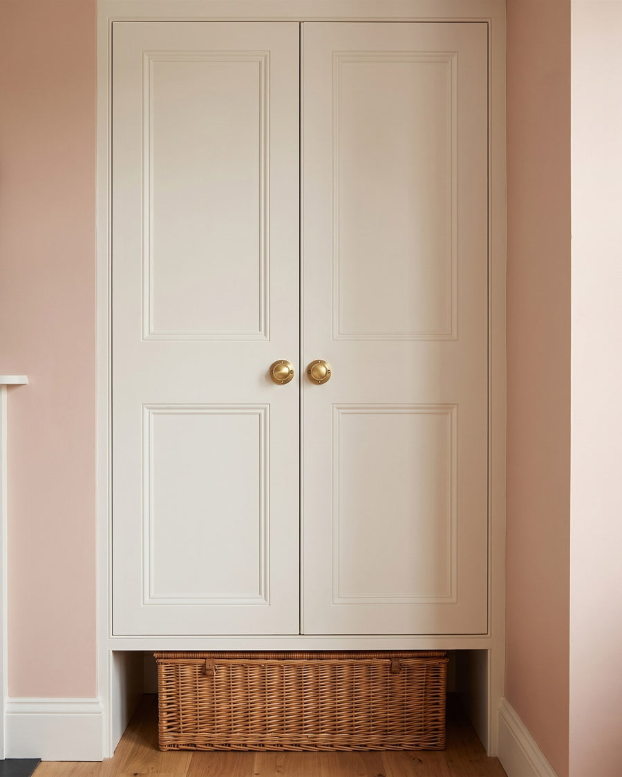 Pair of satin brass Victoria knobs on a warm white panelled wardrobe in a pink alcove, with a wicker hamper beneath