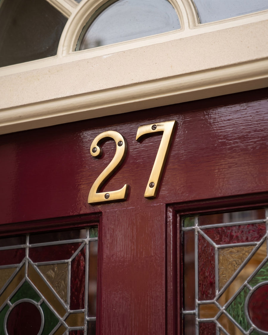Antique brass door numbers 27 on the top rail of an oxblood door above stained glass
