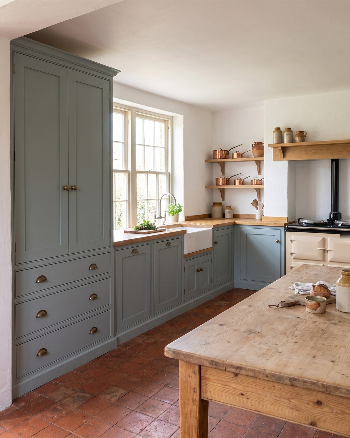 Victorian farmhouse kitchen with blue-grey green in-frame units, oak worktops, a Belfast sink, a cream range cooker, copper pans on oak shelves and a terracotta tiled floor
