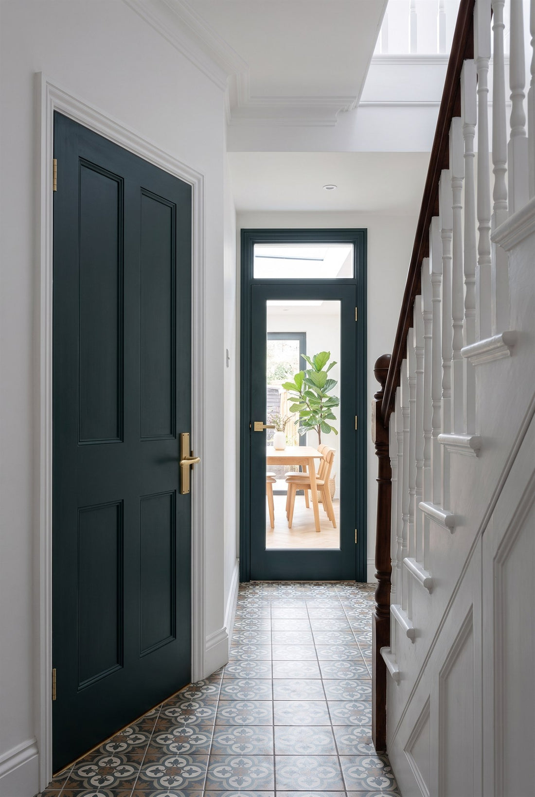 Victorian hall with patterned tiles, a white staircase and an inky green panelled door with a satin brass reeded lever, looking through to a slim glazed door with a satin brass linear lever and a bright extension beyond