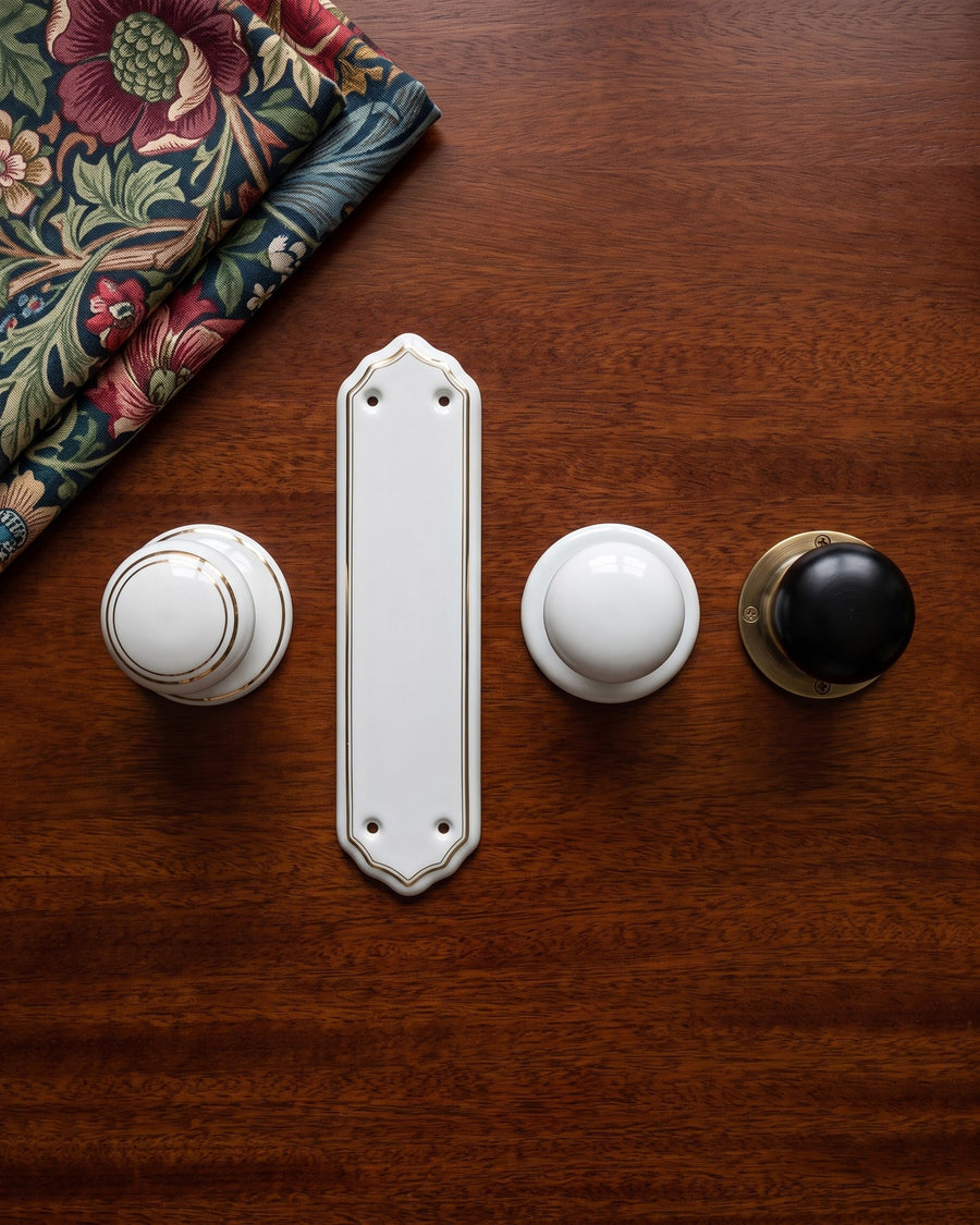 Gold-line porcelain knob and finger plate, plain white porcelain knob and ebony knob on brass laid out on a mahogany table with Morris-style fabric