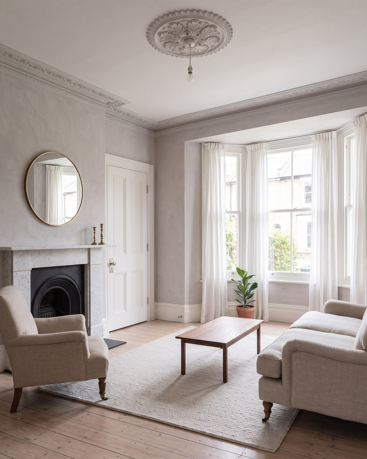 Victorian sitting room with putty limewash walls, a ceiling rose, marble fireplace, linen sofa and armchair and a bay window with sheer curtains