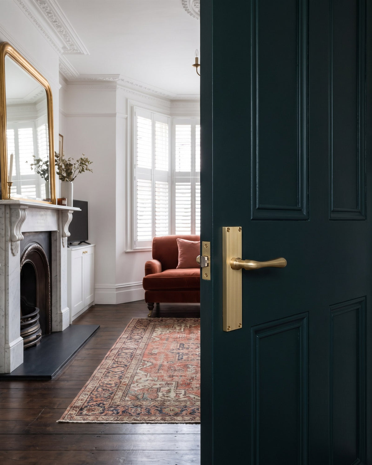 Inky green door with a satin brass Charlbury reeded handle opening onto a Victorian sitting room with a marble fireplace, gilt mirror, shuttered bay and rust velvet sofa