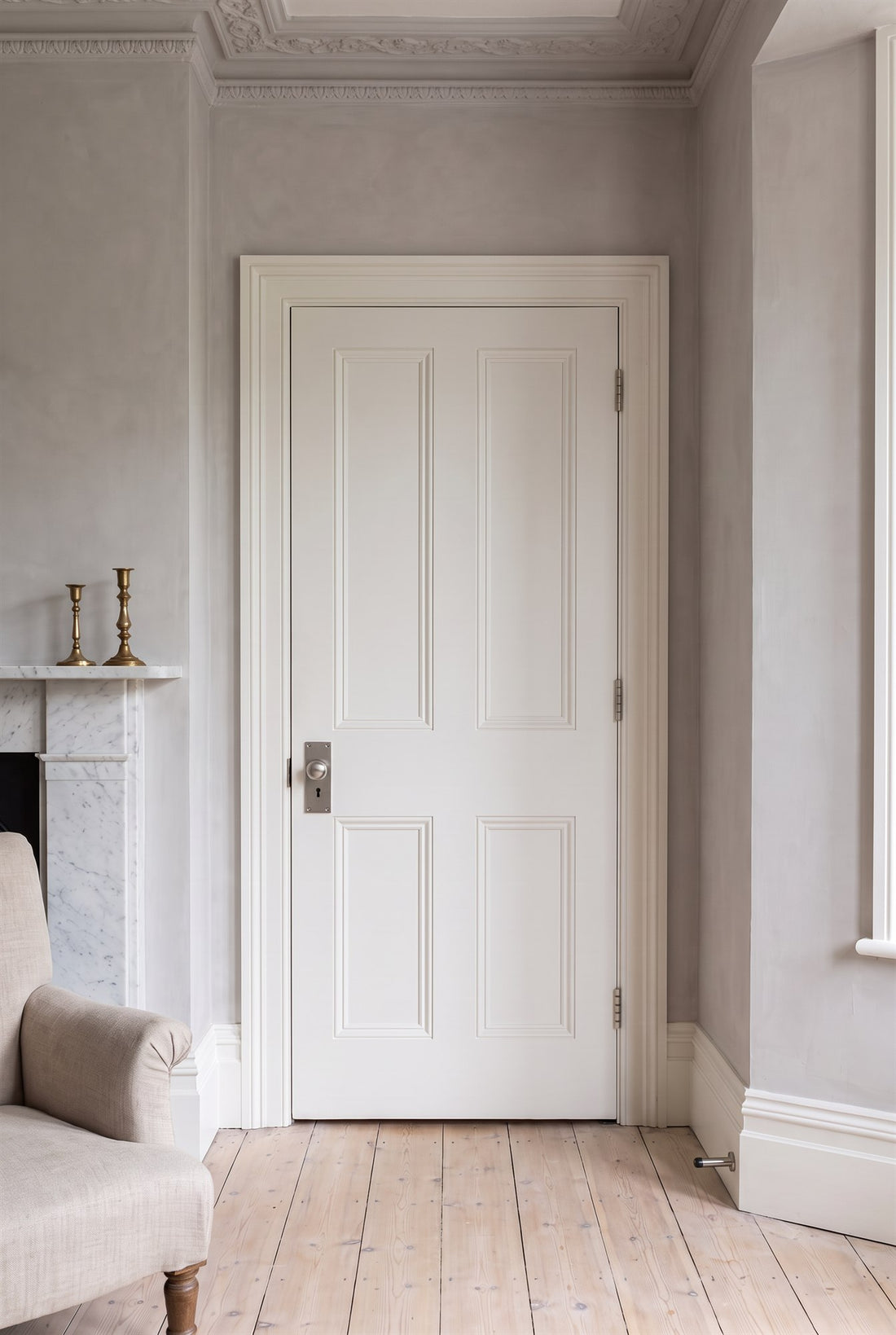 Warm white panelled door in a Victorian sitting room with putty walls and limed pine boards, fitted with a satin nickel knob on lock plate and a cylinder door stop, a marble fireplace beside