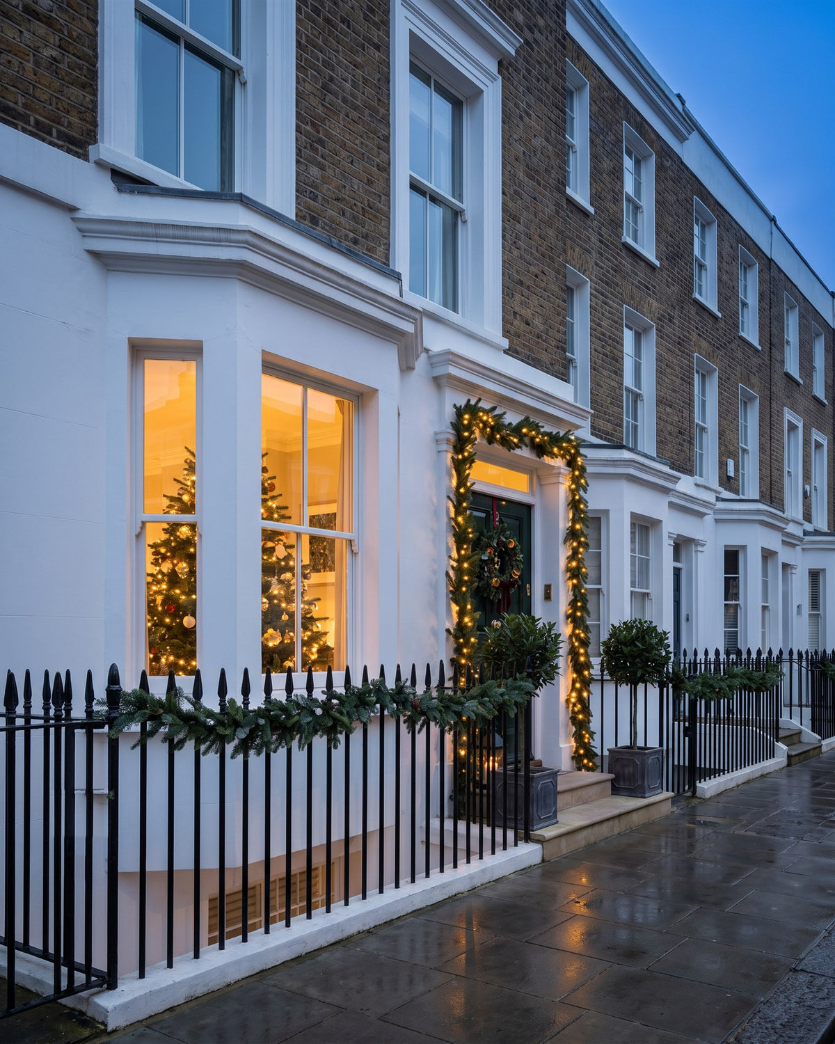 Victorian stucco and brick terrace at dusk with a lit Christmas tree in the bay window, garlanded black railings and a green front door framed by fairy-lit fir garland