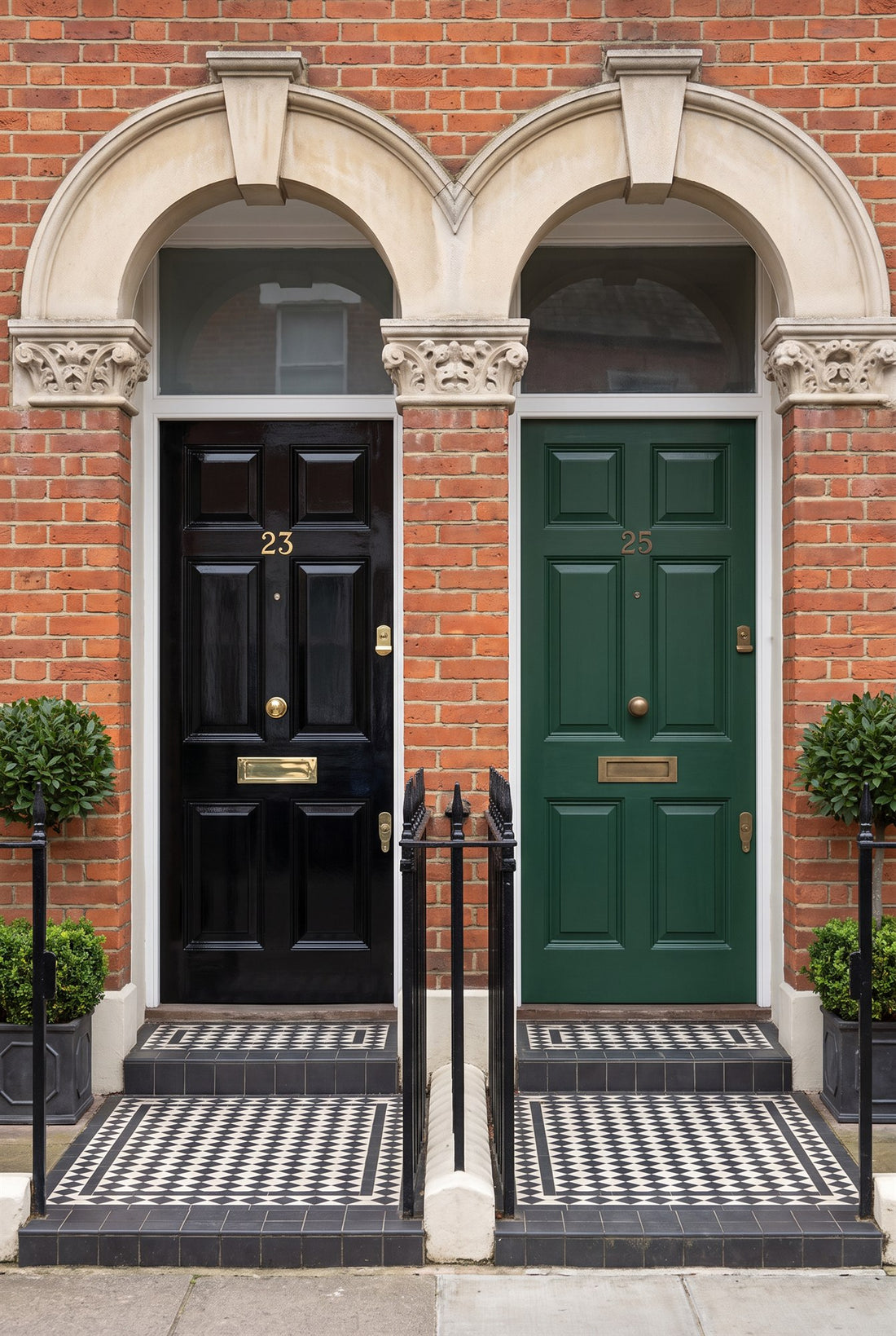 Two neighbouring Victorian terrace front doors, a glossy black door numbered 23 in polished brass and a racing green door numbered 25 in antique brass, under stone arches with tiled steps