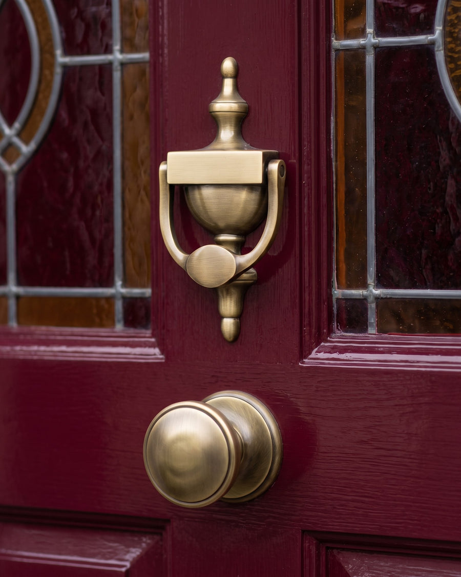 Antique brass Victorian urn knocker above an antique brass centre door knob on an oxblood door between stained-glass panels