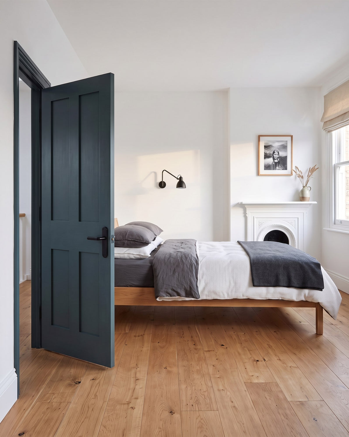 White bedroom with an oak floor, low oak bed with grey and white linen and a white fireplace, a deep green door with a matt black lever standing open