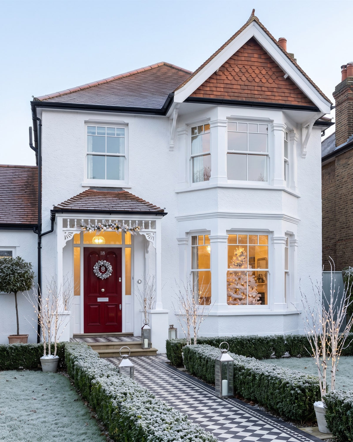 White-rendered Edwardian detached villa on a frosty morning with a red front door in a white fretwork porch, a chequerboard path, frosted box hedges and silver lanterns