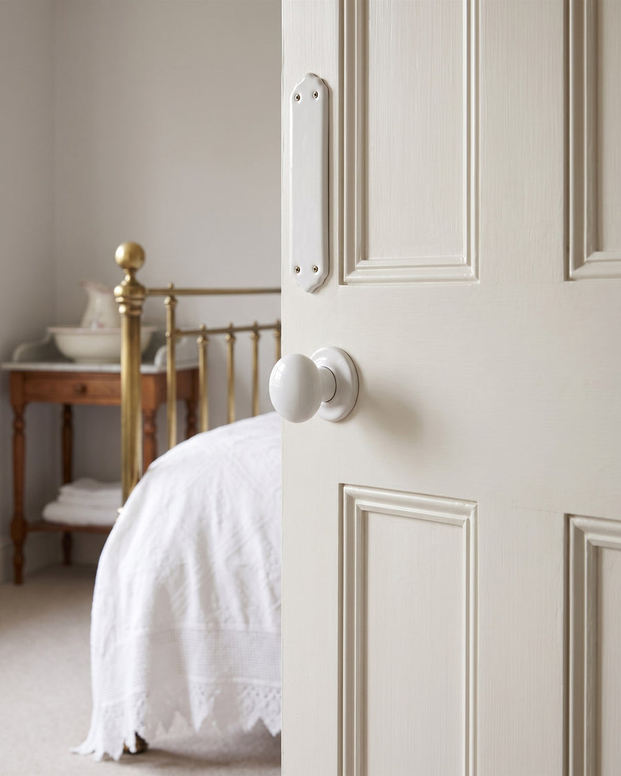 Plain white porcelain knob and finger plate on an off-white bedroom door, brass bedstead and washstand beyond