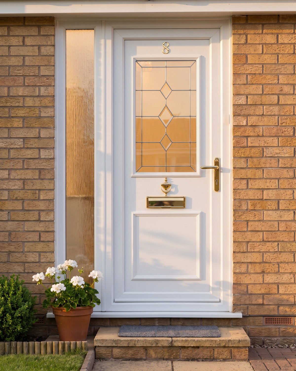 White uPVC front door and side light in warm evening sun, gold number, urn knocker, letter plate and lever glowing against the white